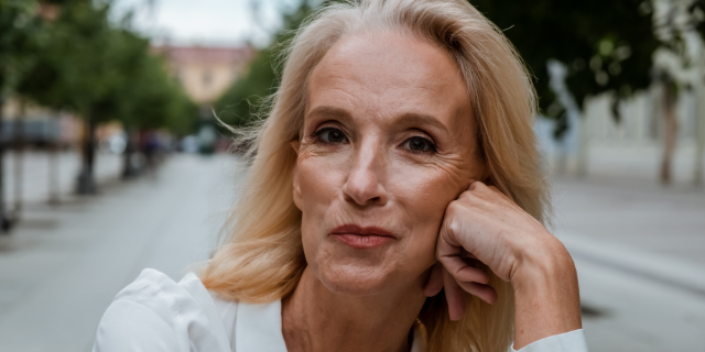 Mulher madura apoia o rosto em uma das mãos. Ela está sentada e o cenário é externo, de uma praça.