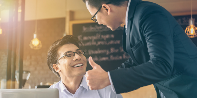 Homem sorri sentado à mesa e olha para cima, onde há outro homem que o olha de volta com aprovação. Este segundo ergue o polegar.