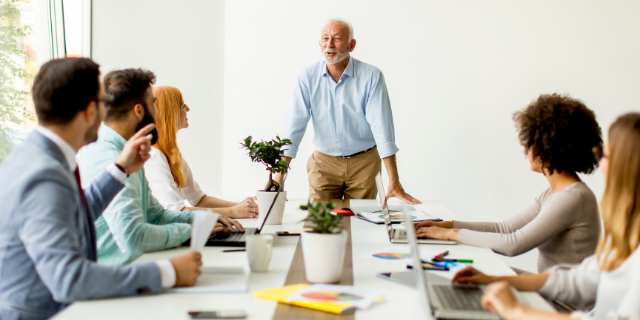 Homem erguido em frente de uma mesa. Abaixo dele, há mais cinco pessoas. O ambiente é de escritório.