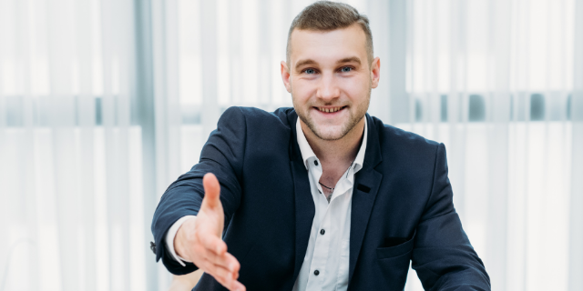 Homem sentado à mesa sorri e estende a mão em direção frontal. O ambiente é de escritório.