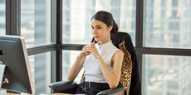 Mulher sentada à mesa com as mãos próximas do rosto. O ambiente é de um escritório.