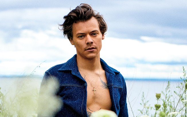 Harry Styles em praia, com camisa jeans de manga comprida aberta, exibindo seu peitoral.