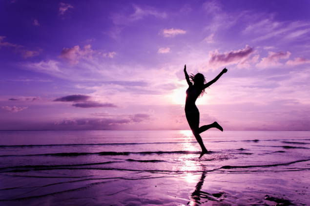 Mulher com as mãos abertas enquanto molha os pés na água da praia. O Sol está se pondo