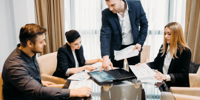 Três pessoas sentadas e um homem de pé enquanto estão em reunião. 