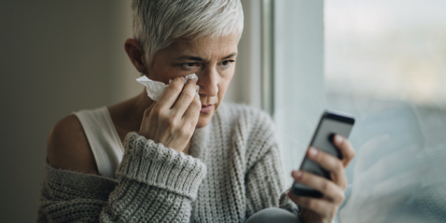 Mulher sentada em um sofá chora ao olhar para celular. Ela enxuga o rosto com lenço de papel branco.