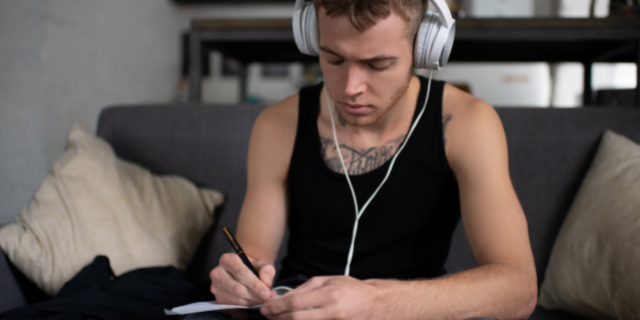 Homem com fone de ouvido branco sentado em sofá. Ele escreve em um caderno sobre sua perna.