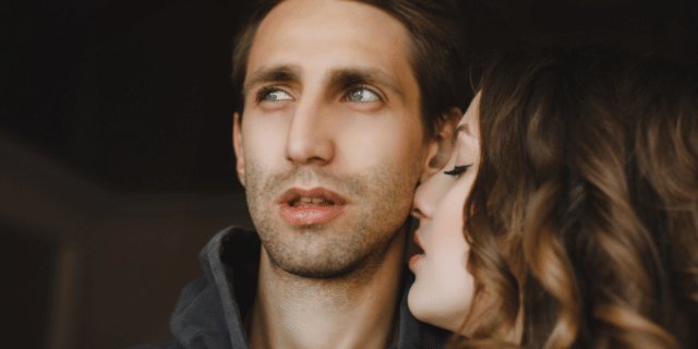 Mulher próxima do pescoço de um homem ao seu lado. Ele olha para o lado oposto ao dela.