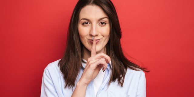 Mulher com o dedo indicador sobre os lábios, indicando silêncio. O fundo é vermelho.