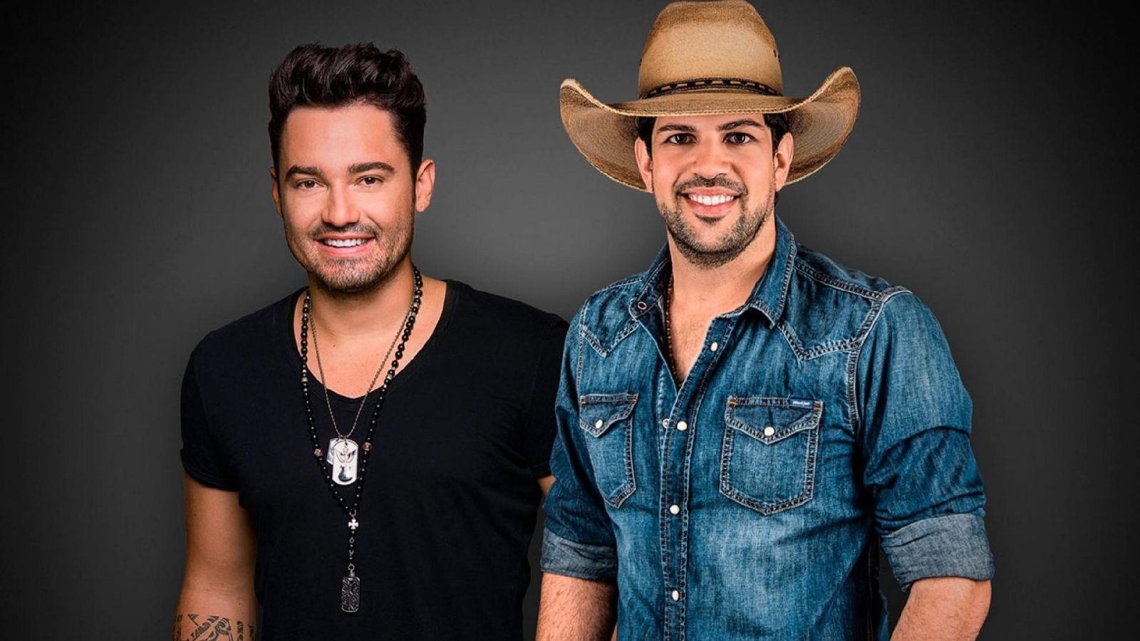 Fernando e Sorocaba sorrindo e olhando para frente em um fundo escuro