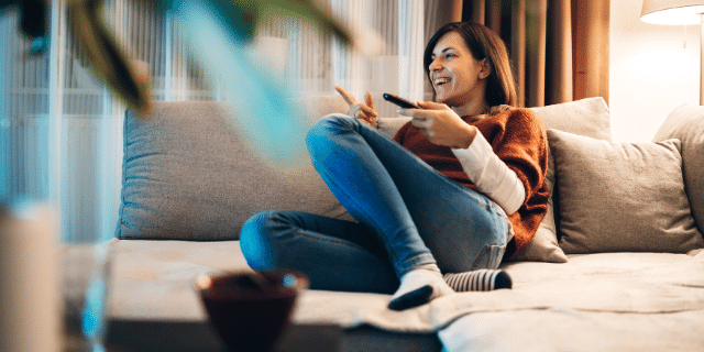 Mulher sentada em sofá sorri enquanto encara um ponto aleatório do ambiente. Ela segura um controle remoto.