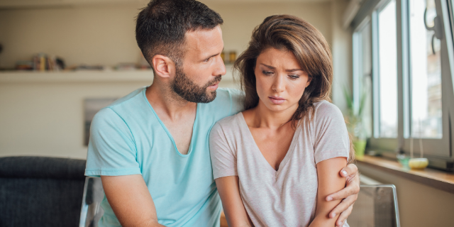 Homem e mulher sentados lado a lado. Ele a abraça e a consola.