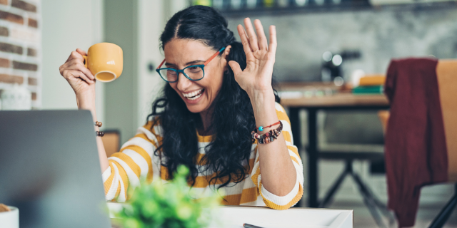 pixelfit / Canva Mulher sentada à mesa sorri ao olhar para a tela do computador. Ela segura uma xícara de porcelana amarela e acena.