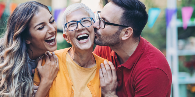Homem e mulher abraçam mulher madura que está em meio aos dois. Os três sorriem.