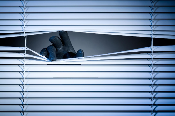 Mão com luvas abrindo uma fresta na persiana de uma janela.