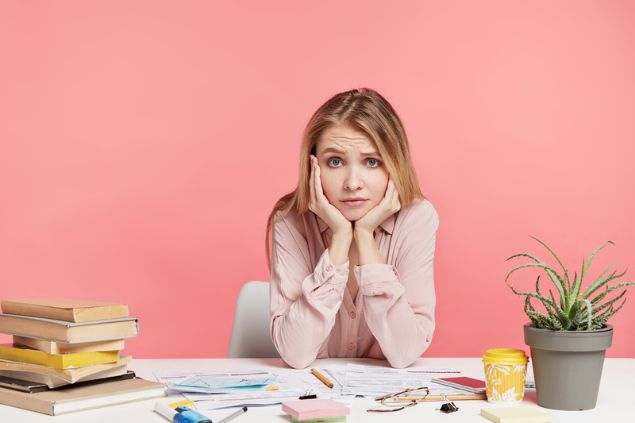 Mulher sentada, com os cotovelos sobre a mesa, apoiando a cabeça nas mãos. Ela está com ar de desalento e cercada por materiais de trabalho/estudo, como livros e papéis.