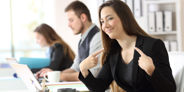 Mulher aponta para si mesma, seu semblante é orgulhoso. Ao fundo, há um homem e uma mulher sentados à mesa enquanto mexem em computadores.