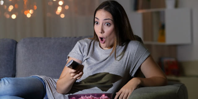 Mulher sentada em sofá com o semblante surpreso e boquiaberta. Ela segura um controle remoto.
