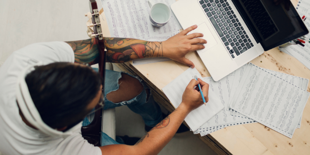 Homem sentado à mesa escreve partituras; sobre a mesa há um computador. Ele está com um instrumento de cordas sobre o colo.