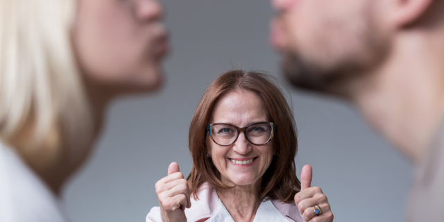 Um casal aproxima os rostos, ao fundo, há uma mulher sinalizando aprovação diante da cena.