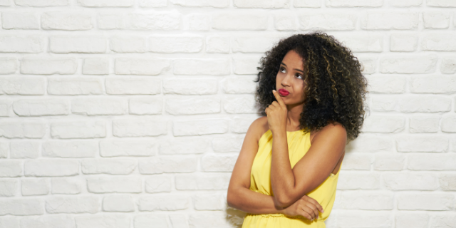 Mulher apoia a mão sobre o queixo. Ela está encostada em uma parede branca e tem o semblante de dúvida.