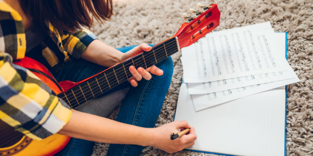 Mulher sentada em tapete escreve em caderno - sobre o caderno, há folhas com partituras. Ela segura um violão próximo ao corpo.