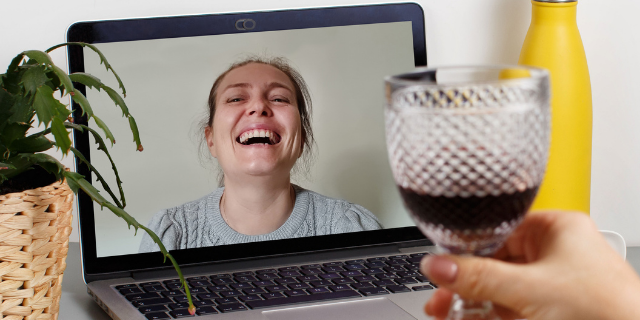 katrinshine / Canva Mulher segura taça de vinho em frente de computador. Na tela, há outra mulher sorrindo. As duas estão em uma vídeo-chamada.