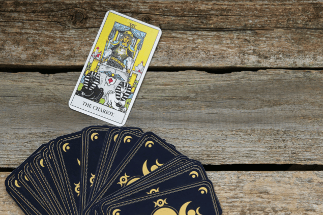 A carta O Carro do Tarot está em destaque sobre uma superfície de madeira. Embaixo, há outras cartas de Tarot viradas para baixo.