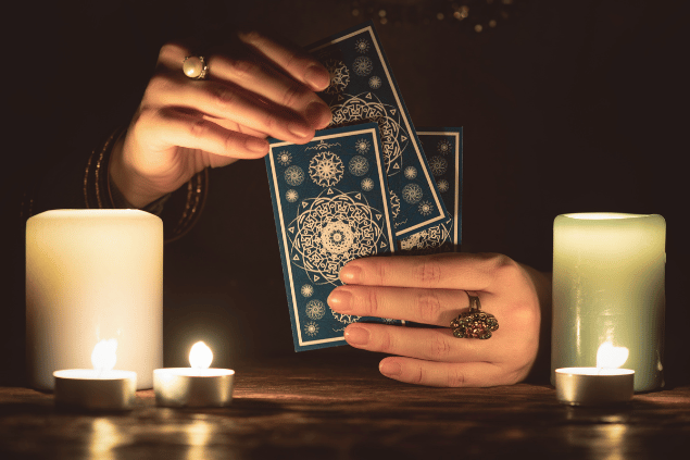 As mãos de uma taróloga segurando três cartas de Tarot estão em destaque na imagem. Ao lado, há algumas velas.