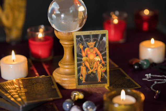 A carta O Diabo do Tarot está em pé sobre uma mesa. Ela está apoiada em uma bola de cristal. Há um baralho com outras cartas de Tarot e velas ao lado.