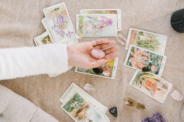 A mão de uma taróloga segurando um cristal está em destaque na imagem. Em cima de uma mesa, há várias cartas de Tarot e outros cristais.