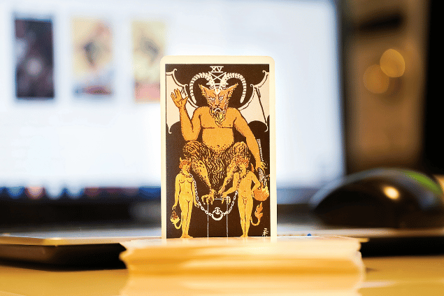 Na imagem, há a carta O Diabo do Tarot na posição normal e em pé. Ao fundo, há um notebook aberto.