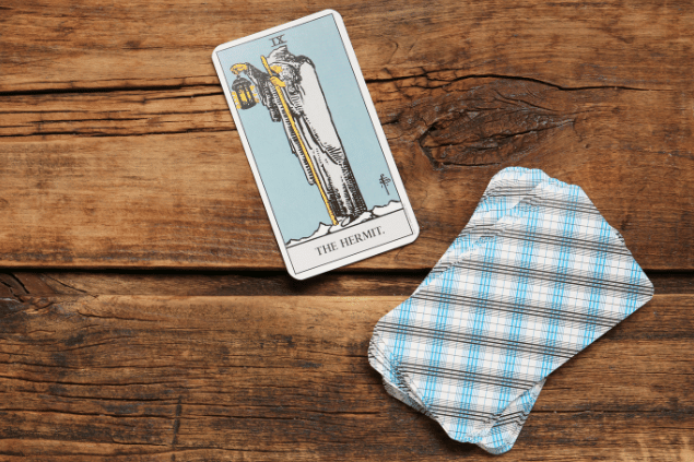 A carta O Eremita do Tarot está em destaque sobre uma superfície de madeira. Embaixo, há outras cartas de Tarot viradas para baixo.