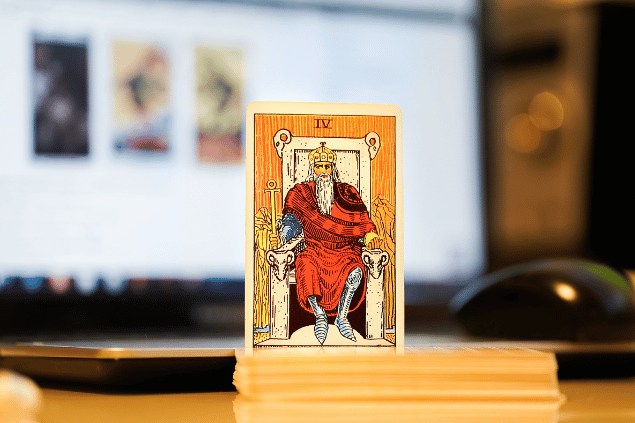 Na imagem, há a carta O Imperador do Tarot na posição normal e em pé. Ao fundo, há um notebook aberto.