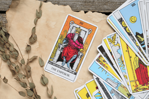 A carta O Imperador do Tarot está em destaque sobre uma superfície de madeira e de um papel. Ao lado, há outras cartas de Tarot.