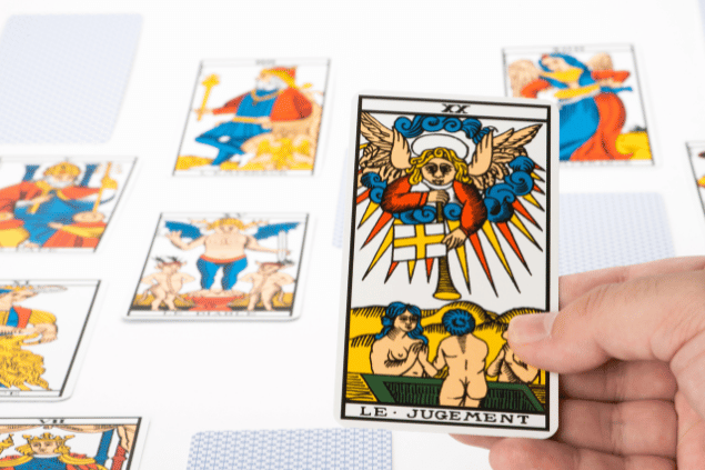 A mão de uma pessoa segurando a carta O Julgamento do Tarot está em destaque na imagem. Ao fundo, há outras cartas dispostas sobre uma mesa.