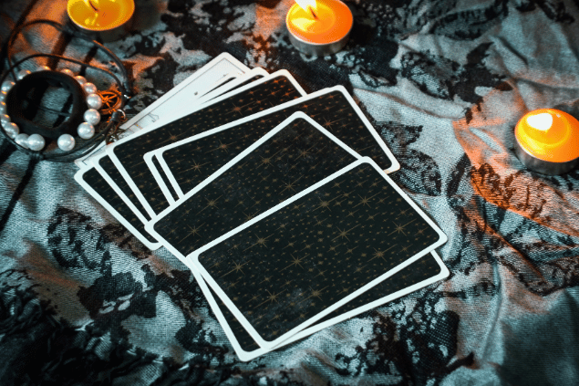 Várias cartas de Tarot estão espalhadas e viradas para baixo sobre uma mesa. Ao lado, há velas acesas.