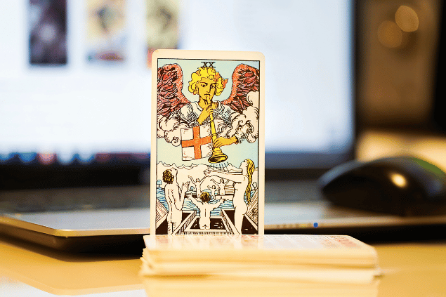 Na imagem, há a carta O Julgamento do Tarot na posição normal e em pé. Ao fundo, há um notebook aberto.
