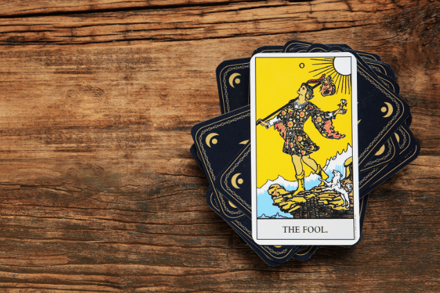 A carta O Louco do Tarot está em destaque sobre uma superfície de madeira. Embaixo, há outras cartas de Tarot viradas para baixo.