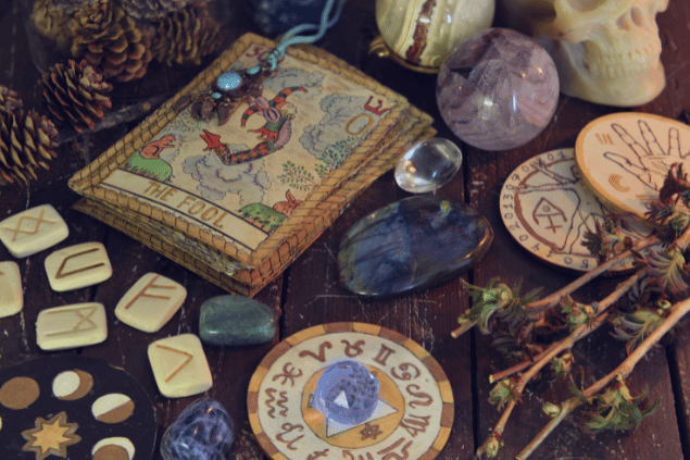 A carta O Louco do Tarot está em destaque sobre uma mesa de madeira. Ao lado, há runas, flores e cristais.