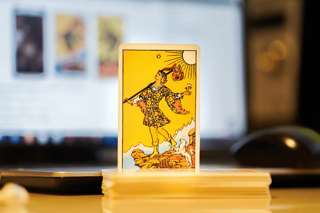 Na imagem, há a carta O Louco do Tarot na posição normal e em pé. Ao fundo, há um notebook aberto.