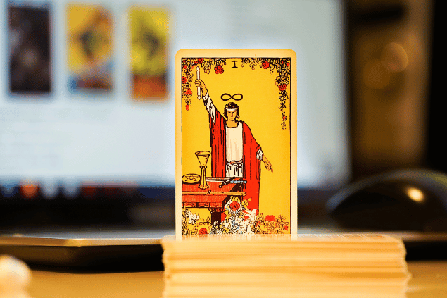 Na imagem, há a carta O Mago do Tarot na posição normal e em pé. Ao fundo, há um notebook aberto.