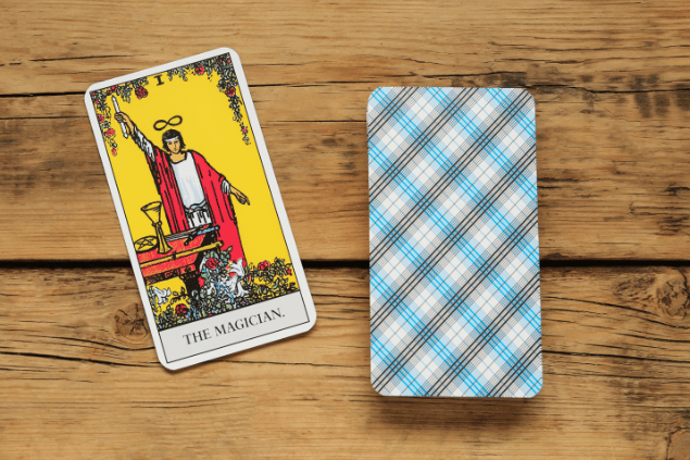 Na imagem, há a carta O Mago sobre uma mesa de madeira. Ao lado, um baralho de cartas de Tarot está virado para baixo.