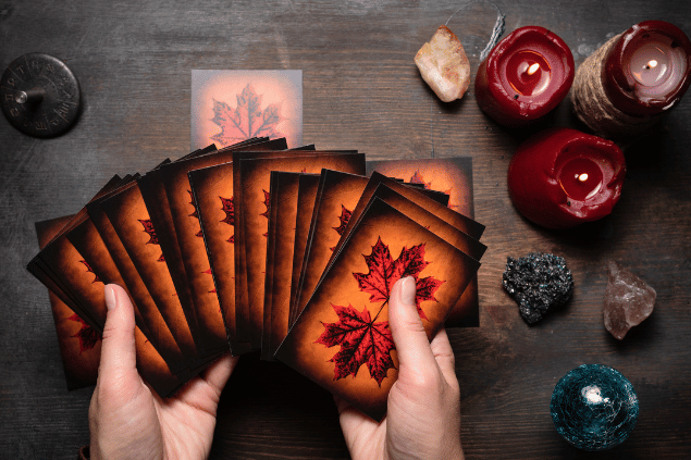 As mãos de uma taróloga segurando várias cartas de Tarot viradas para baixo estão em destaque na imagem. Algumas velas e cristais estão em cima de uma mesa.