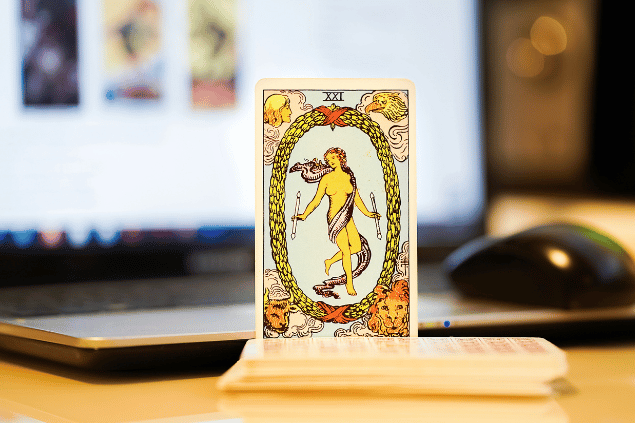 Na imagem, há a carta O Mundo do Tarot na posição normal e em pé. Ao fundo, há um notebook aberto.
