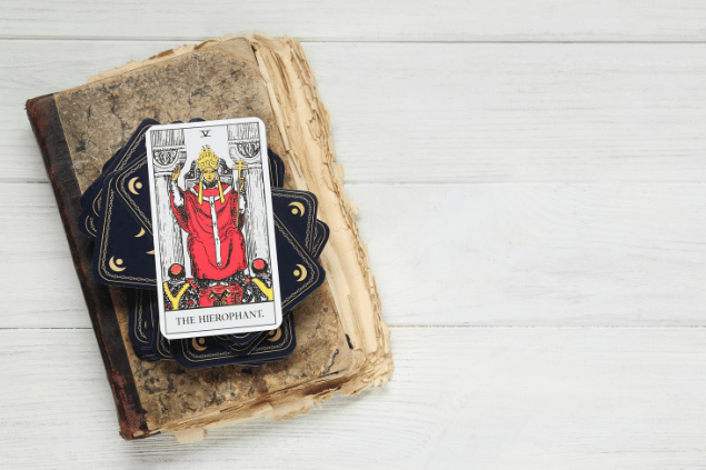 A carta O Papa do Tarot está em destaque sobre um baralho com outras cartas viradas para baixo e sobre um livro antigo. 