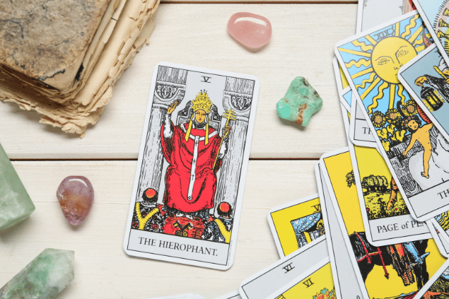 A carta O Papa do Tarot está em destaque sobre uma superfície de madeira branca. Ao lado, há outras cartas de Tarot e cristais.