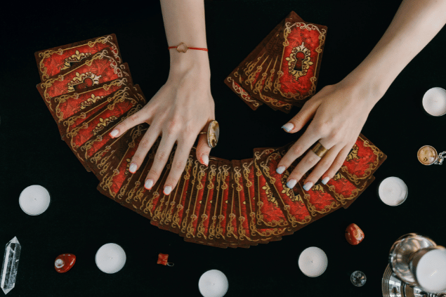 As mãos de uma taróloga sobre várias cartas de Tarot em sequência estão em destaque na imagem. Ao lado, há velas, cristais e pingentes.