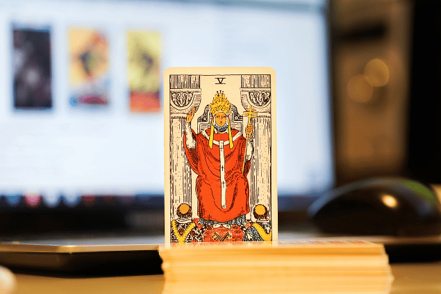 Na imagem, há a carta O Papa do Tarot na posição normal e em pé. Ao fundo, há um notebook aberto.
