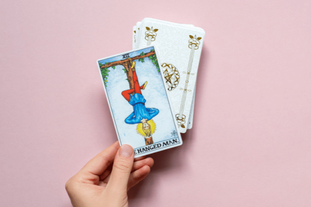 A mão de uma pessoa segura a carta O Pendurado do Tarot que está em destaque na imagem. Embaixo dela, há um baralho com outras cartas viradas para baixo.