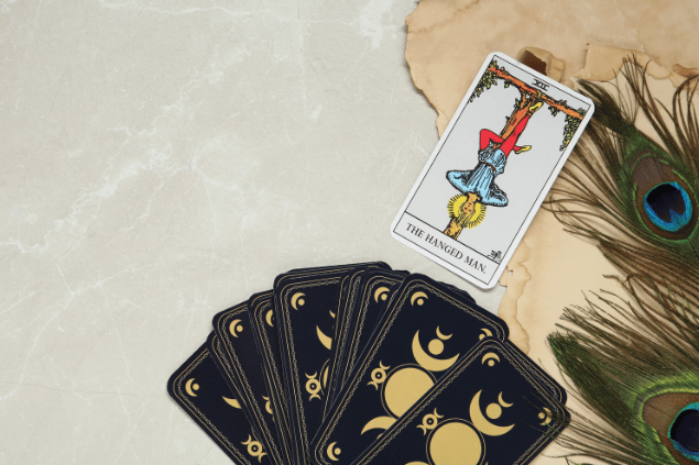A carta O Pendurado do Tarot está em destaque sobre uma mesa e sobre um papel. Embaixo, há outras cartas de Tarot.